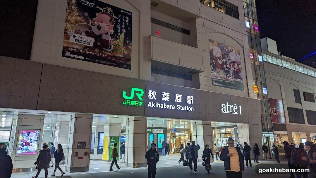 Night view of Akihabara Station entrance with visible signage including 'JR東日本' and 'akihabara Station'. Adjacent storefronts display illuminated signs like 'atre1', anime-themed posters, and digital billboards. Pedestrians walk along the sidewalk in front.
