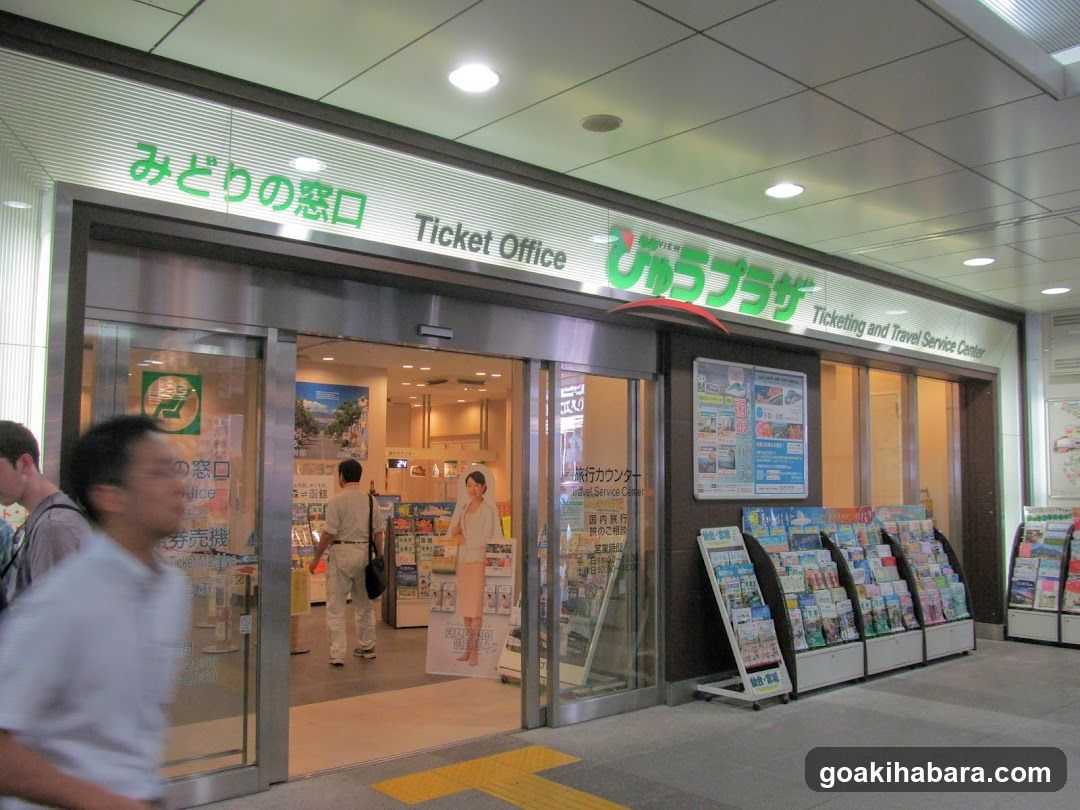 A Japanese train station ticket office with signage reading 'Midori no Madoguchi' and 'Ticket Office'. The entrance features glass doors, and there are racks of brochures outside.