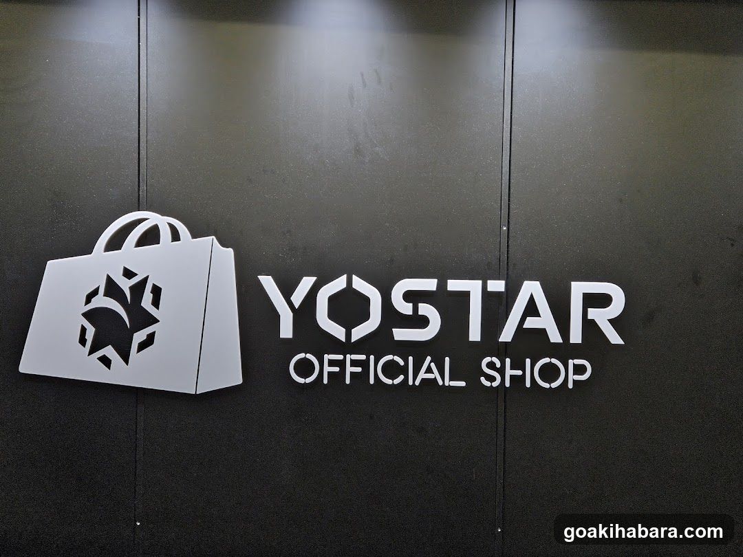 A close-up photo of a shop entrance featuring a metallic sign on a dark wall. The sign displays the word 'YOSTAR' in bold, stylized letters with a shopping bag icon to the left containing a geometric design. Below 'YOSTAR', the words 'OFFICIAL SHOP' are visible.