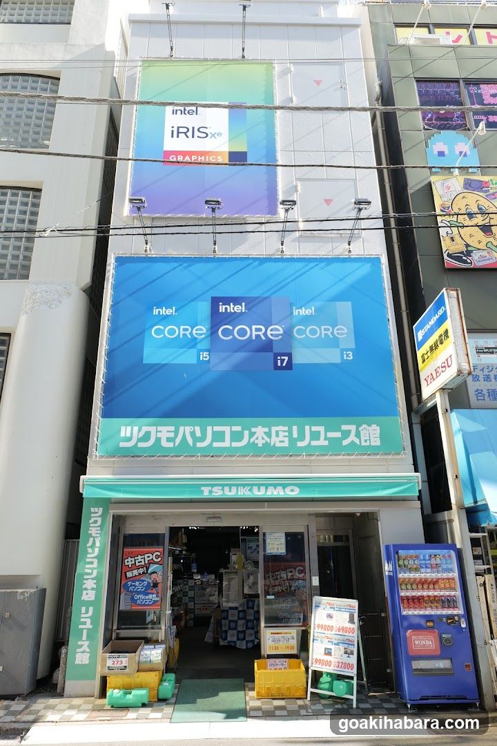 A street-level view of a computer store named TSUKUMO, featuring large Intel graphics advertisements above the entrance. The storefront includes signage in Japanese and English, with visible product displays inside.