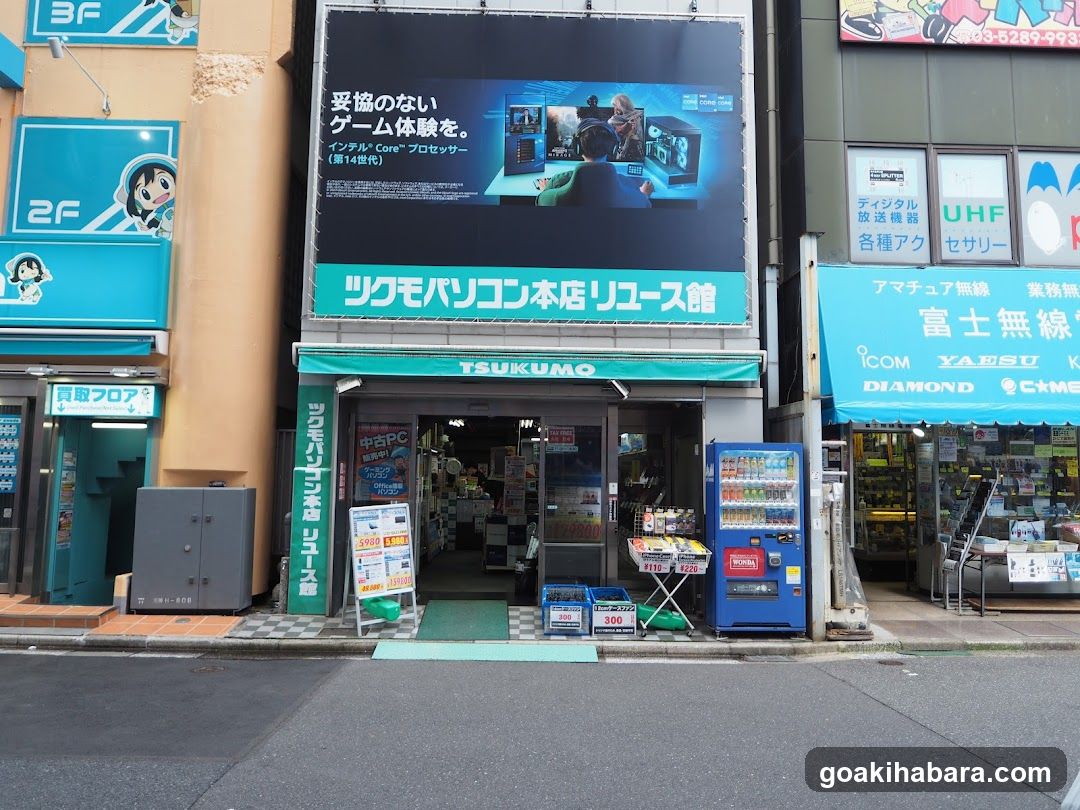 A street-level view of a TSUKUMO electronics store entrance, featuring signage in Japanese and English, with visible vending machines and promotional displays outside the shop.