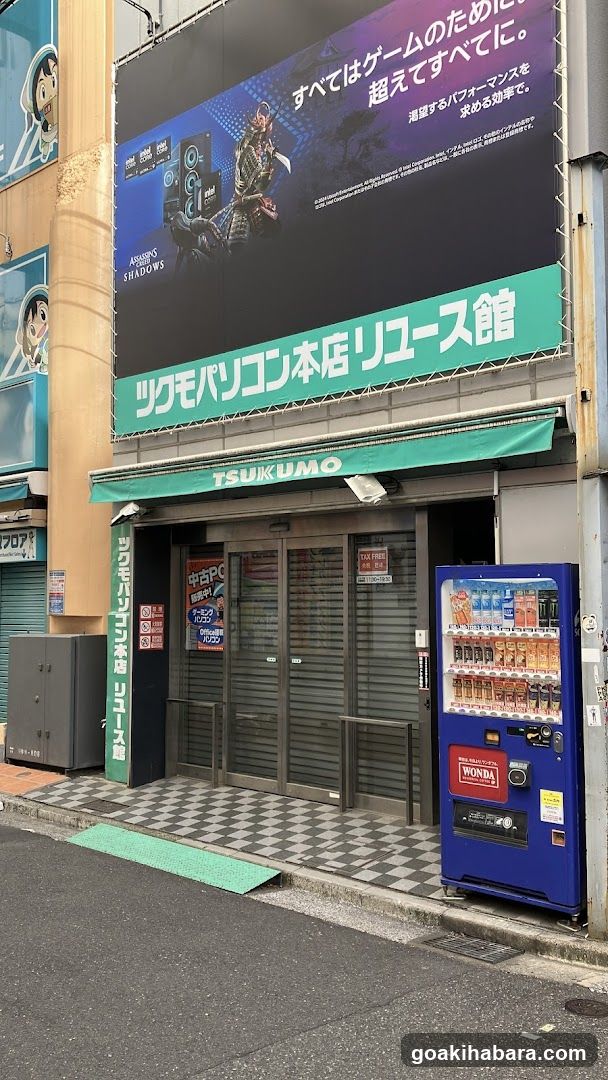 A shop front with a large billboard advertising 'STADIUM W5' and Japanese text above the entrance. The store sign reads 'TSUKUMO' in English and Japanese characters on a green awning. A blue vending machine stands to the right of the entrance, displaying various drinks.