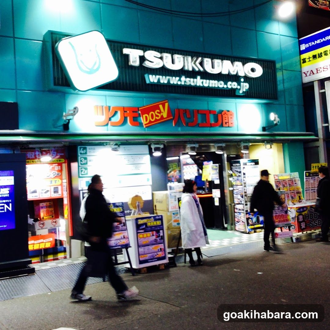 Night view of TSUKUMO building exterior with illuminated signage, entrance doors, and promotional displays outside.