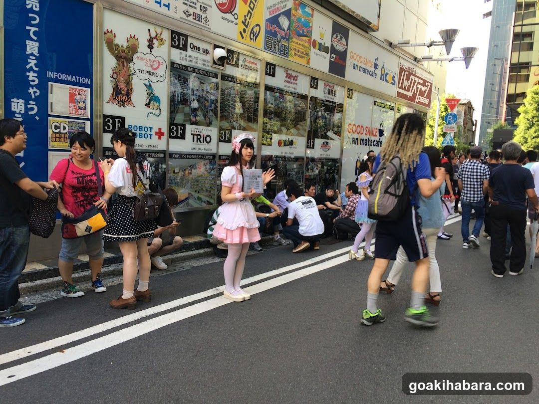 A busy street scene featuring a shop front with visible signage, including '3F ROBOT BOT', 'TRIO', and 'GOOD SMILE an matcafe'. Several people are gathered outside the store, some standing and others sitting on the curb. The shop facade displays various posters and advertisements.