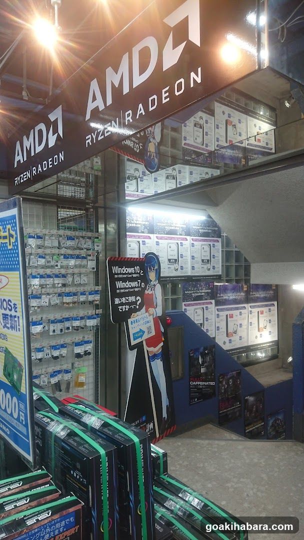 A storefront entrance featuring a large AMD and Ryzen Radeon sign above the door. Inside, shelves display various products with blue packaging. A poster promotes Windows 10 upgrade services next to an anime character illustration. Stacks of boxed merchandise are arranged near the entrance.