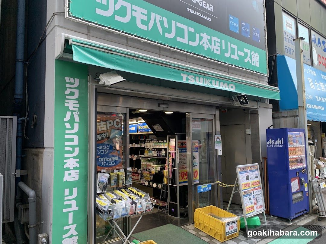 The image shows the exterior of a TSUKUMO reuse shop, featuring green signage with Japanese and English text. The entrance is open, revealing shelves inside stocked with various items. A blue vending machine labeled 'Asahi Soft Drinks' stands to the right. Outside displays include promotional posters, price lists, and a yellow crate filled with products. The storefront has glass doors and windows, allowing visibility into the well-lit interior.