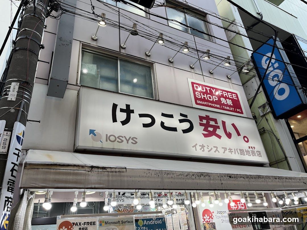 A street-level view of a shop front with signage reading 'けっこう安い' (Keikou Anai) and 'DUTY FREE SHOP'. The sign also mentions 'IOSYS' and lists products like smartphone, tablet, PC. A blue banner with white text is visible on the right side.