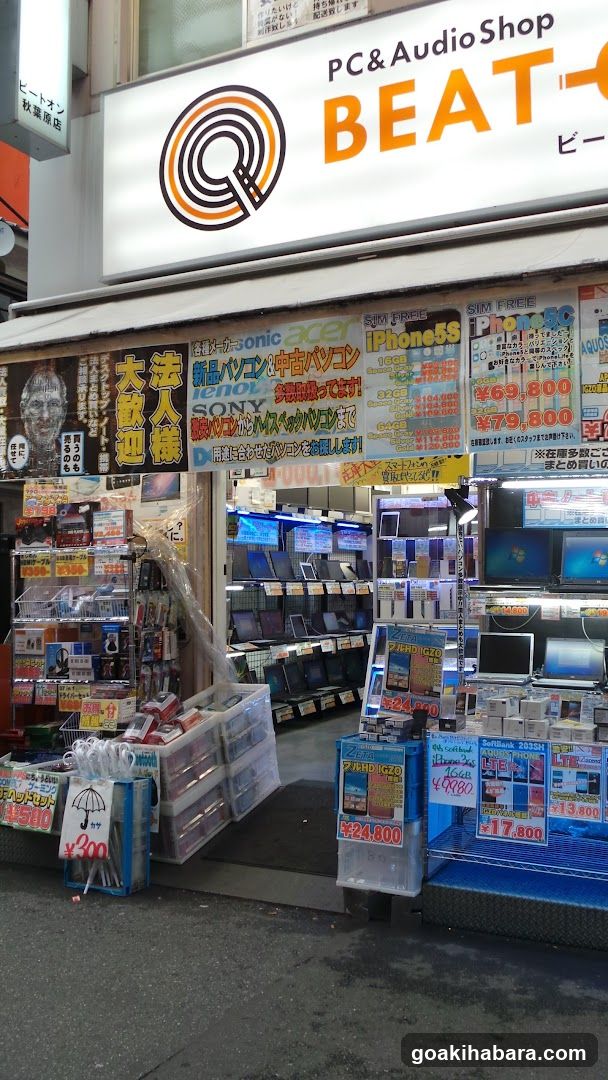 A storefront of a shop named BEAT, specializing in PCs and audio equipment. The signboard is prominently displayed above the entrance with the name 'BEAT' and 'PC & Audio Shop'. Below the main sign, there are various advertisements and price lists for products like iPhones, laptops, and other electronics. The entrance is open, revealing shelves stocked with items inside. There are also promotional signs and displays outside the store.