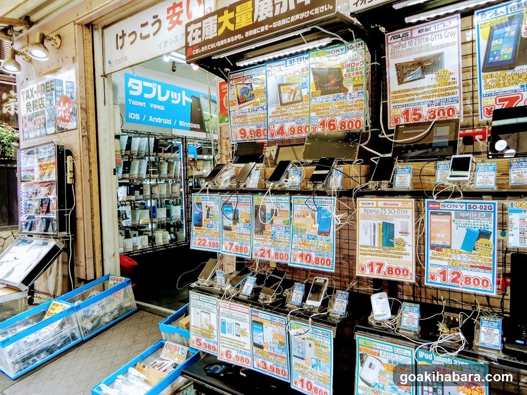 A brightly lit electronics storefront with hanging price tags and product displays. Visible signage includes Japanese characters and English labels like 'Tablet', 'iOS / Android / Windows'. Multiple posters advertise devices such as Xperia phones, iPads, and tablets with prices in yen (e.g., ¥15,800, ¥7,980). A blue sign reads 'タブレット' (tablet) above a glass display case filled with gadgets. The entrance is open, revealing shelves inside.