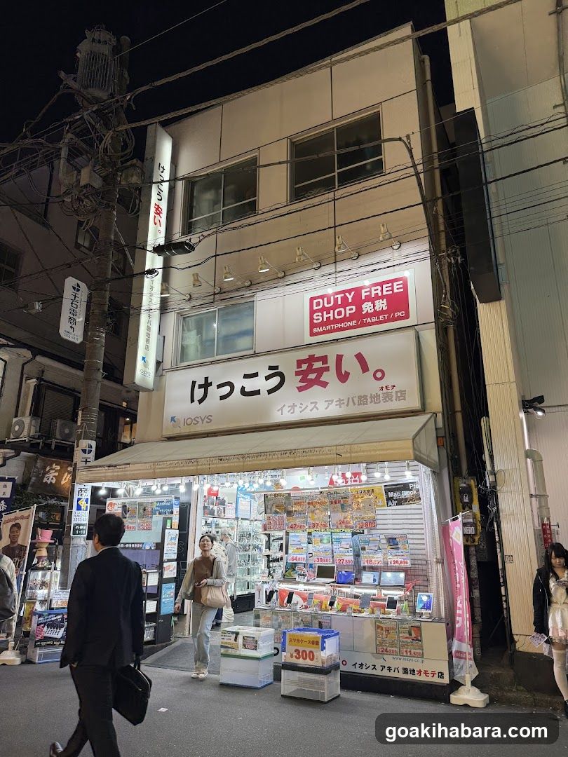 Night view of a two-story shop with 'DUTY FREE SHOP' and Japanese signage, displaying electronics like smartphones/tablets under bright lights.