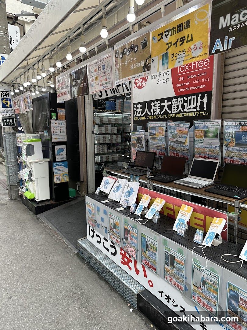 The image shows the exterior of an electronics store with multiple signs and advertisements. Visible items include laptops on display, promotional posters, and a sign reading '法人様大歓迎' (Welcome to businesses). The shop front features various electronic products like computers and accessories.