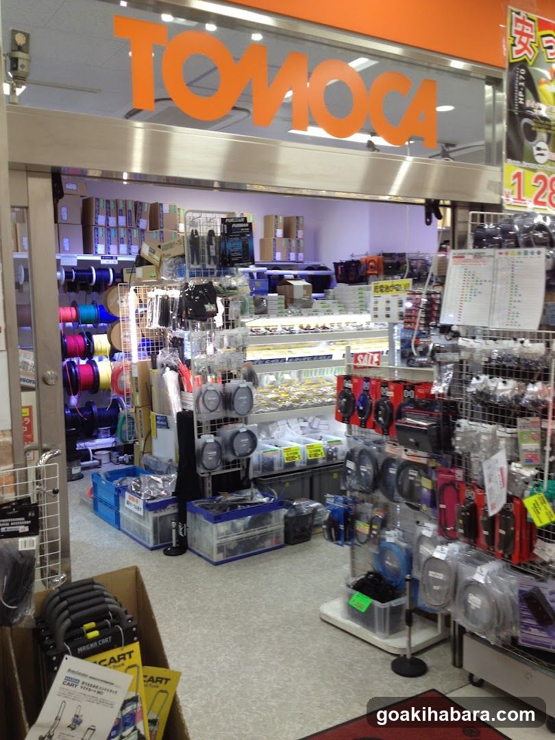 The image shows the entrance of a TOMOCA store, with its orange signage prominently displayed above the doorway. Inside, various products are visible, including spools of thread in different colors, packaged items on shelves, and promotional materials. The interior is well-lit, showcasing an array of merchandise neatly arranged.