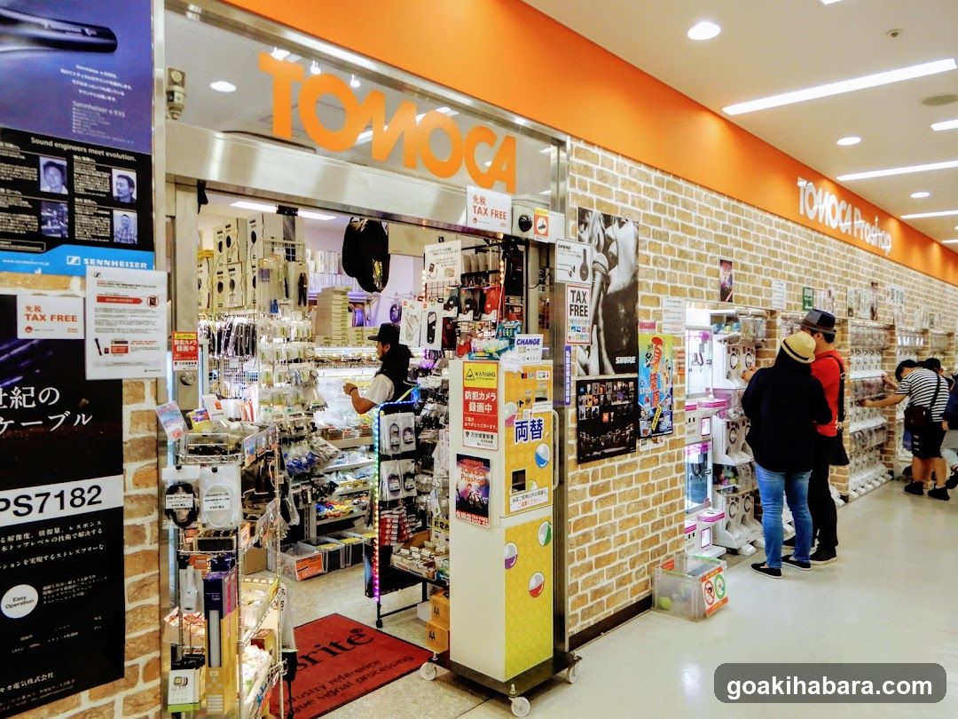 Interior shot of TOMOCA electronics store showing entrance, brick walls, product displays, and customers browsing. Signage visible above door.