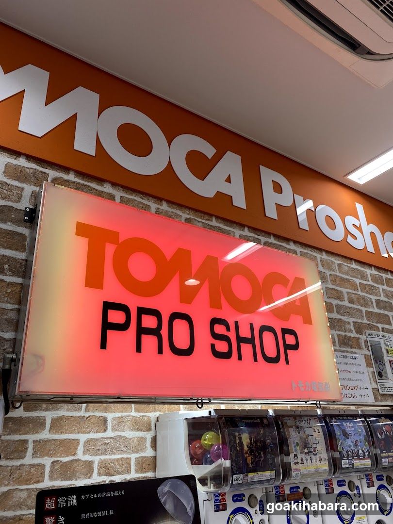The image shows the storefront of a shop named TOMOCA PRO SHOP. The sign is prominently displayed with 'TOMOCA' in orange and 'PRO SHOP' in black on a glowing background. Above it, part of another sign reading 'MOCA Prosh...' is visible against an orange background. Below the main sign, there are several gashapon machines displaying various items.