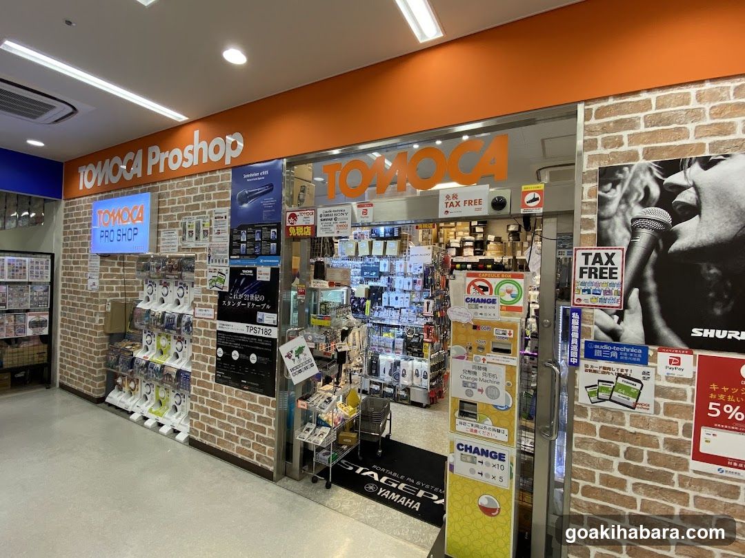 Storefront facade with brick walls and orange signage reading TOMOCA Proshop. Entrance displays various electronics products, including headphones and accessories. Posters advertise tax-free shopping and currency exchange services. Yamaha branding visible on floor mat.