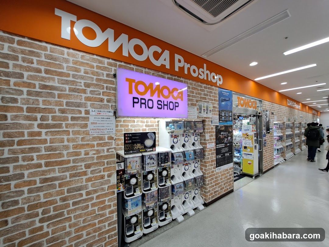 The image shows the exterior of a TOMOCA Proshop store, featuring brick-patterned walls and an orange sign with white lettering. A purple neon sign reading 'TOMOCA PRO SHOP' is mounted on the wall. Several vending machines are lined up along the storefront, displaying various products. The interior is visible through glass doors, showing shelves stocked with merchandise.