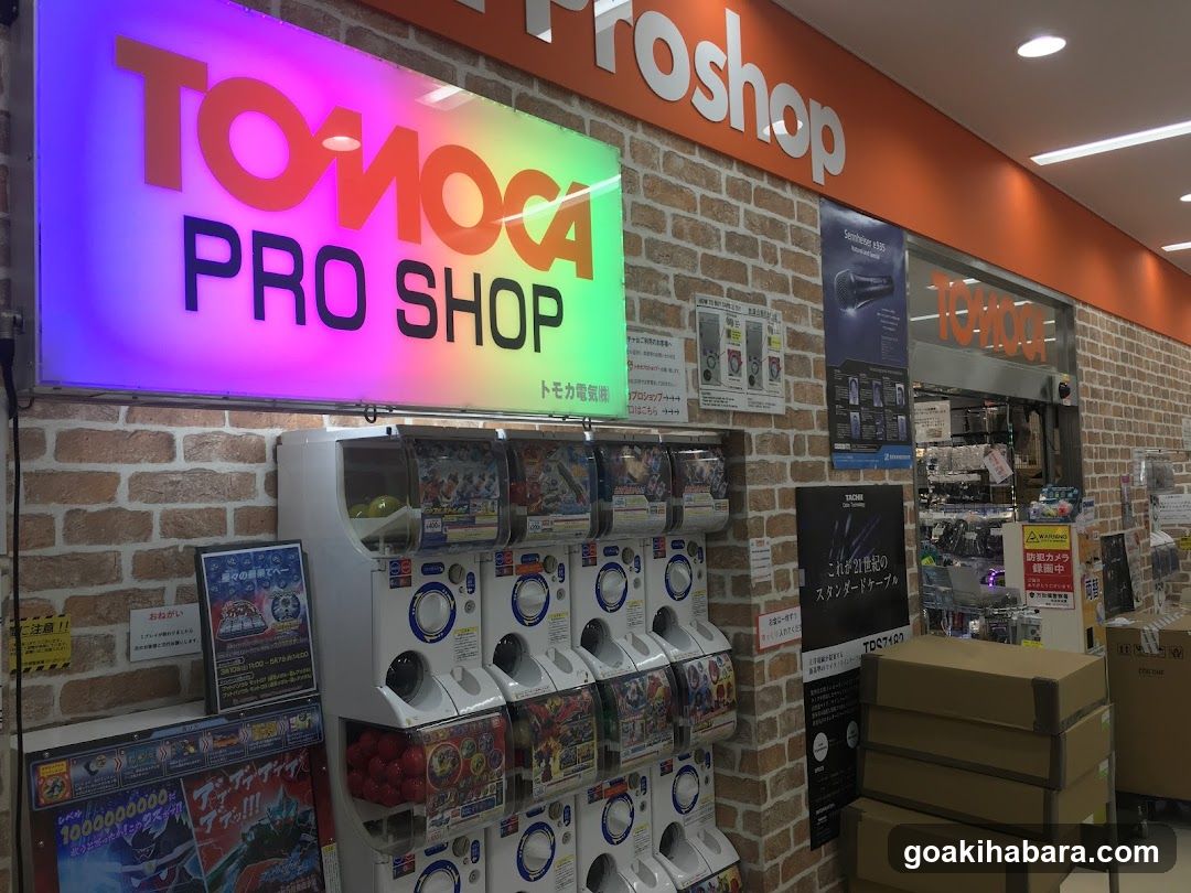 Storefront with brick wall facade, illuminated 'TOMOCA PRO SHOP' sign above entrance, multiple gachapon machines displaying colorful capsules, promotional posters on walls, stacked cardboard boxes near entrance, and visible interior shelves stocked with merchandise.
