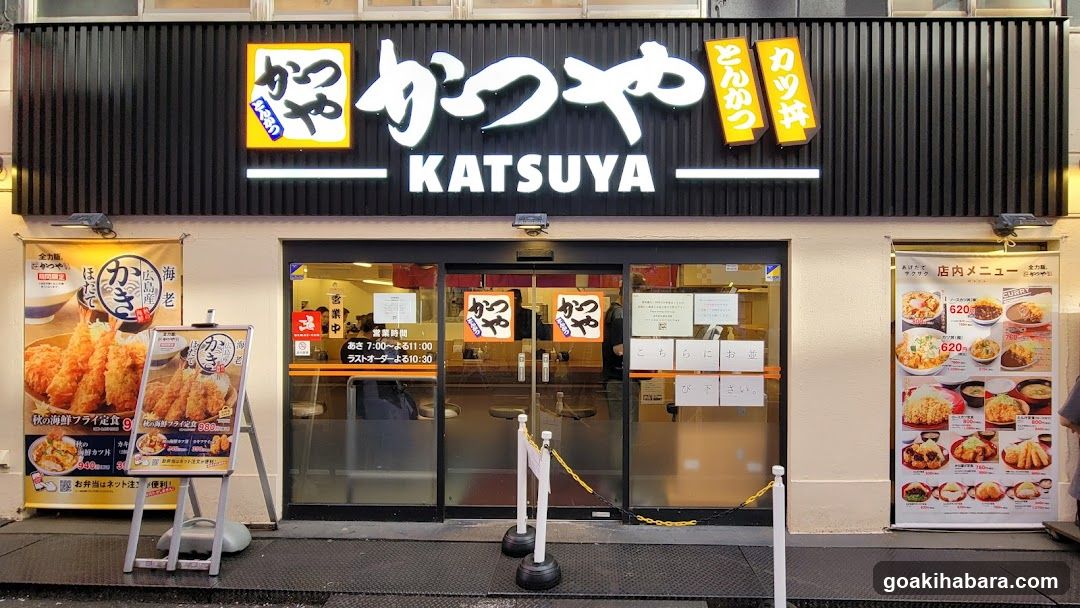 The image shows the exterior of a Katsuya restaurant, featuring its prominent signage with Japanese and English text. The entrance is marked by glass doors with notices posted on them. A menu board stands to the left, displaying various food items like tempura and fried fish. To the right, another poster lists inside menu options with prices.