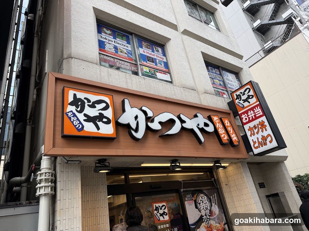 A street-level view of a Katsu-ya restaurant entrance, featuring prominent signage with Japanese characters and English text indicating 'Tonkatsu' and 'Fried Chicken'. The building facade includes windows displaying advertisements for Tokyo Electric Power and other services. A vertical signboard lists menu items like Tonkatsu and Takoyaki.