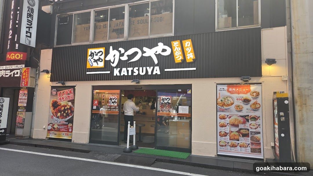 A street-level view of a Katsuya restaurant storefront featuring prominent signage with Japanese and English text, glass doors leading to the interior, and illuminated menu boards on either side displaying food items.