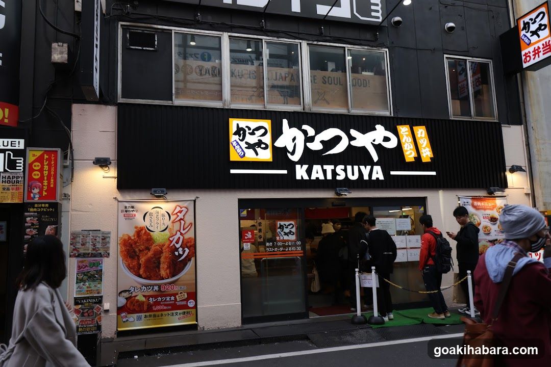 A street-level view of a Katsuya restaurant entrance with customers queuing at the door. The black facade features illuminated signage reading 'KATSUYA' and Japanese characters, along with vertical banners promoting fried chicken products. A large poster on the left wall displays a crispy fried chicken dish with pricing details visible below it.