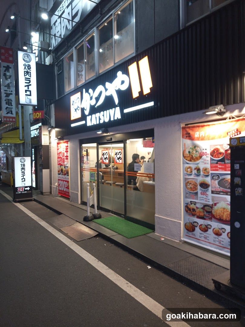 The image shows the exterior of a Katsuya shop, featuring illuminated signage with Japanese and English text. The entrance is visible through glass doors, with staff inside. A menu board displays various food items with prices.