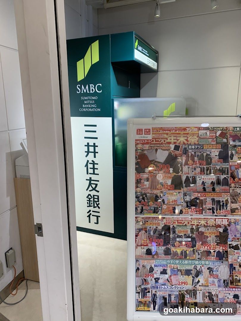 Photo of the entrance to a Sumitomo Mitsui Banking Corporation (SMBC) branch, featuring signage with Japanese characters and English text. A promotional display board is visible in the foreground.