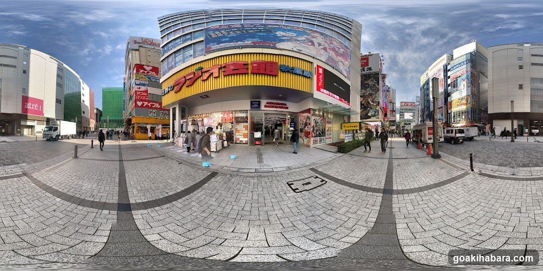 A wide-angle view of a multi-story shopping center with prominent yellow and red signage featuring Japanese characters. The entrance is bustling with pedestrians, and various advertisements are visible on the building's facade.