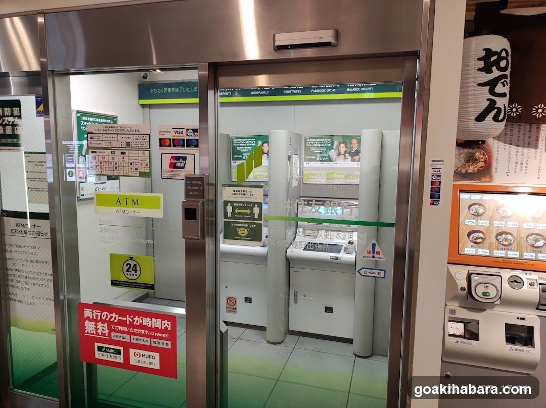 Photo of a bank ATM entrance with glass doors and signage indicating 24-hour service. Visible signs include 'ATM', '24 hours', and Japanese text about card usage. A menu board is mounted on the right wall displaying food items with prices.