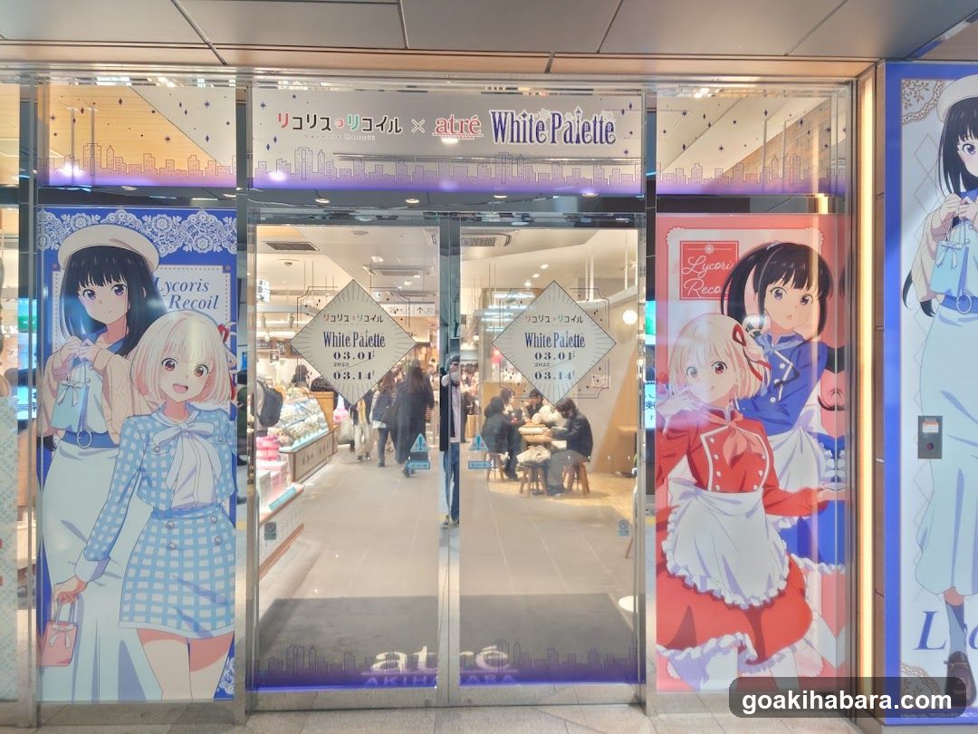 A storefront entrance featuring large anime-style character posters on both sides of the glass doors. The central signage reads 'リコリス・リコイル x atré White Palette'. Inside, customers are seated at tables in a brightly lit cafe area with dessert displays visible.