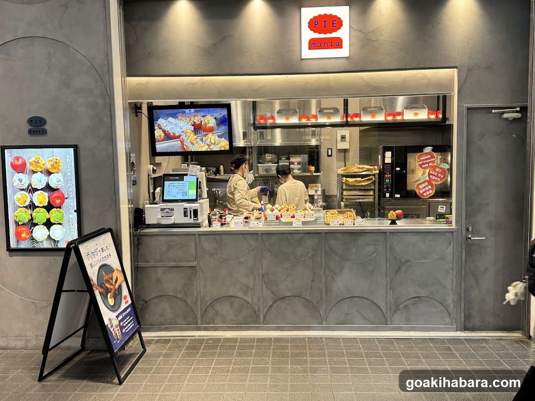 A modern storefront with a gray facade and arched design elements. The shop features a large service window where staff are preparing food. Above the counter, there is a sign reading 'PIE mania' in red and white. To the left of the entrance, a digital menu board displays colorful pie options. A freestanding poster on the sidewalk advertises a promotion or special offer.