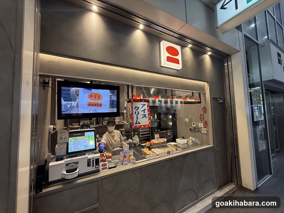A modern shop front with a large glass window displaying food items and staff inside. A sign above the entrance reads 'PIE MANZO' in Japanese characters. Inside, there is a TV screen showing promotional content, a cash register, and various food products on display.