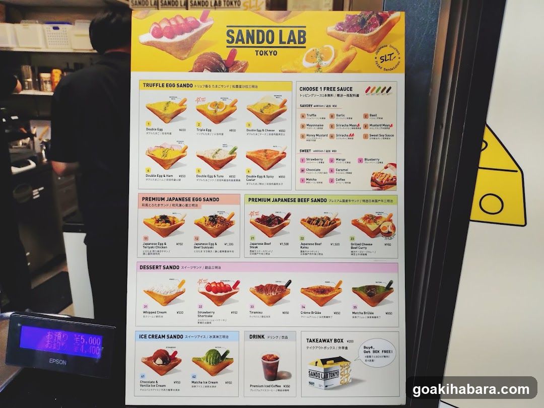 A vertical menu board for Sando Lab Tokyo, featuring various egg sandwich options like Truffle Egg Sando and Premium Japanese Beef Sando. The menu includes sections for sauces, desserts, ice cream sandwiches, drinks, and a takeaway box offer.