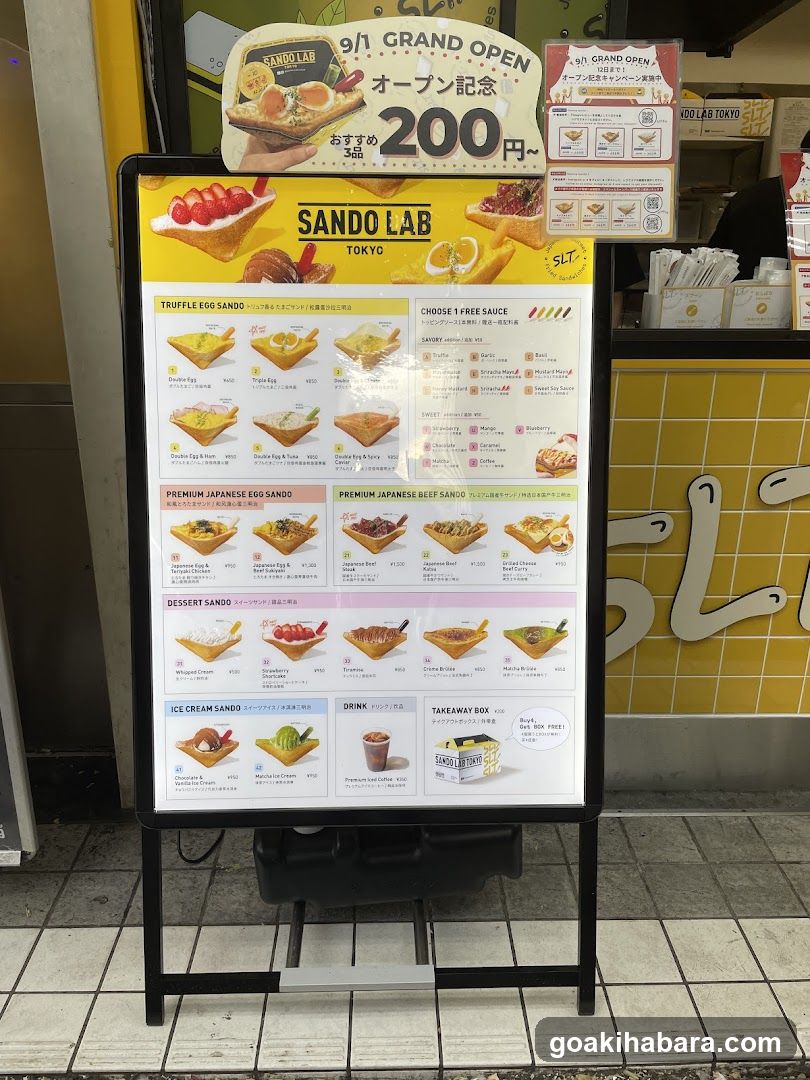 A large outdoor menu board displaying various sandwich options from Sando Lab in Tokyo, including Truffle Egg Sando, Premium Japanese Egg Sando, Dessert Sando, Ice Cream Sando, and Takeaway Box.