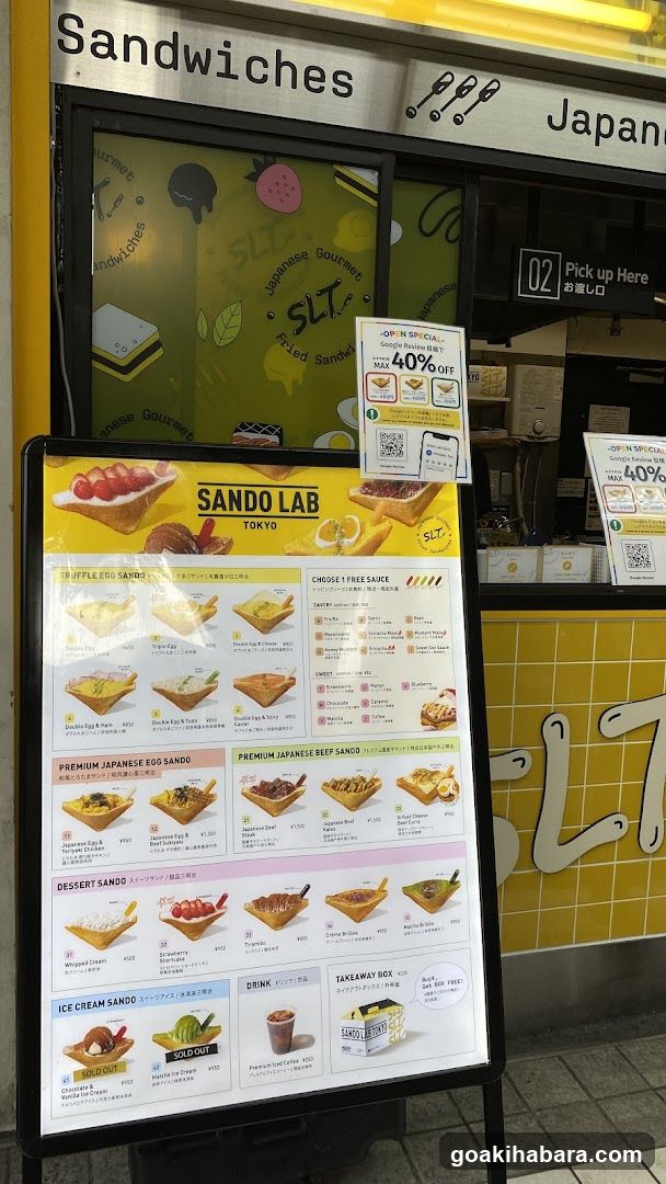 A large outdoor menu board displaying various sandwich options from Sando Lab Tokyo, including Jiffle Egg Sandwich, Premium Japanese Egg Sando, Beef Sando, and Ice Cream Sando with prices in yen.