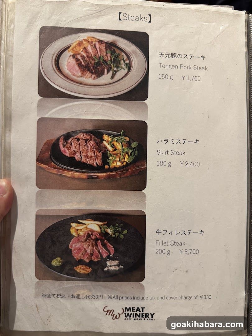 A close-up of a printed menu page featuring three steak dishes with photos, Japanese names, English translations, weights, and prices in yen. The bottom includes tax/cover charge info and the restaurant logo 'MW MEAT WINERY'.
