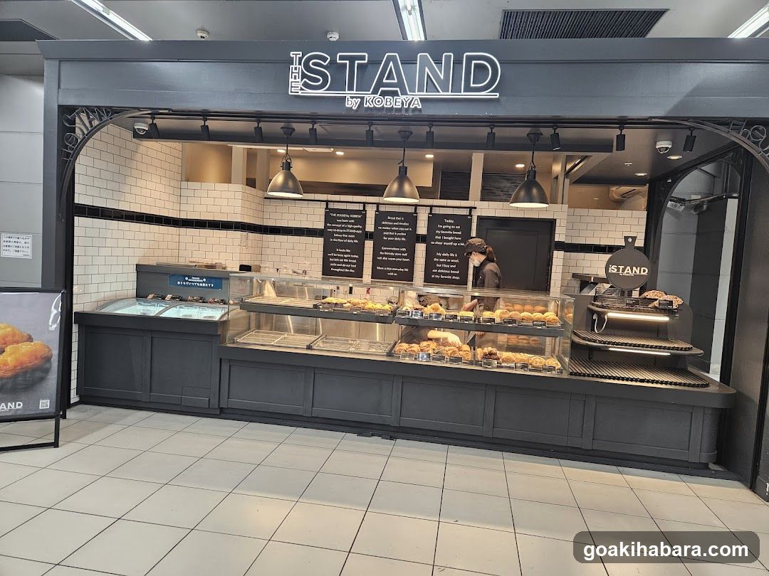 A modern bakery storefront with a dark gray facade and white subway tile backsplash. The sign 'THE ISLAND by KOBeya' is prominently displayed above the entrance. Inside, there are glass display cases filled with various baked goods like breads, pastries, and sandwiches. A staff member is visible behind the counter. Lighting fixtures hang from the ceiling, illuminating the interior.