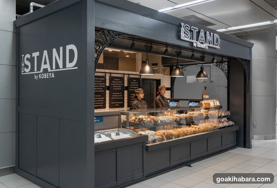 A modern dark gray storefront with 'THE ISLAND by KOBAYA' signage on the facade and above the entrance. Inside, two staff members stand behind a glass display case filled with assorted baked goods like croissants and pastries. Menu boards are visible in the background.