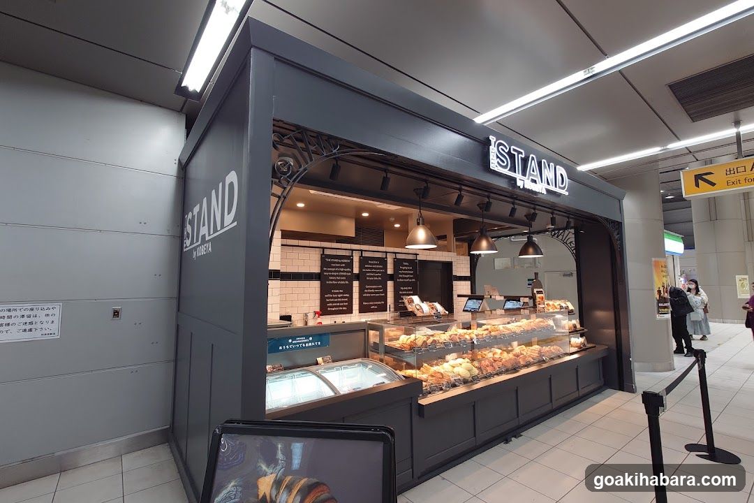 A modern dark gray kiosk with 'ISTAND' signage on the facade and interior display case filled with pastries. Interior shows menu boards, cash registers, and hanging lights.
