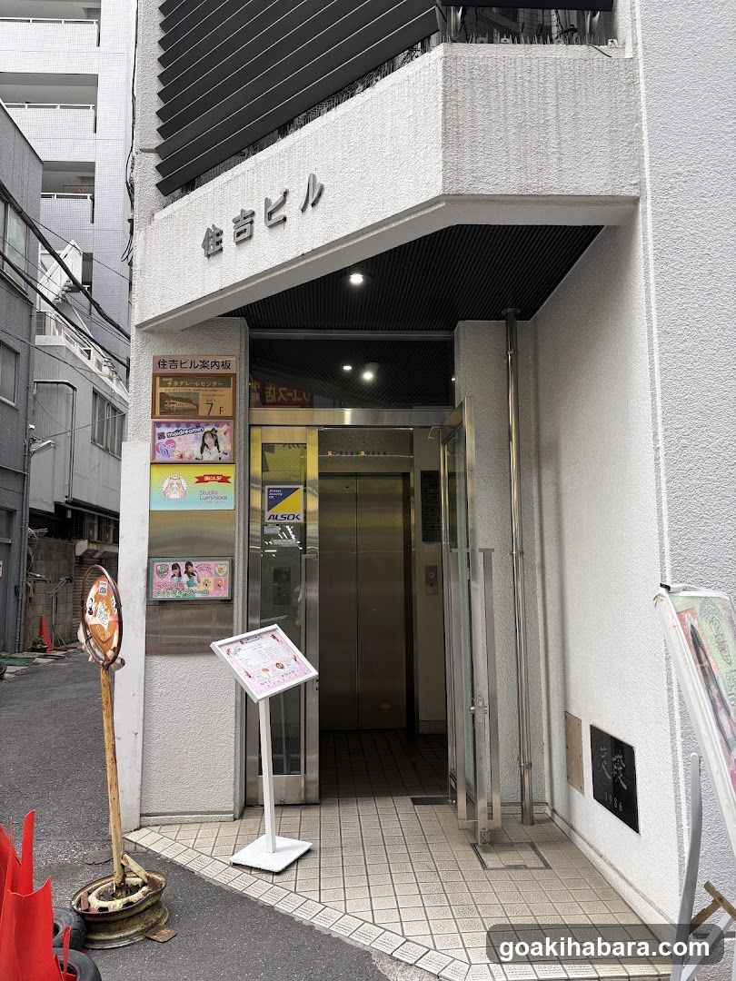 The image shows the entrance to a building, likely an apartment or office complex, featuring a glass door leading into an elevator shaft. Above the entrance, there is Japanese text on the facade. To the left of the entrance, several posters are displayed, including one mentioning '7F' and another with colorful illustrations. A signboard stands in front of the entrance, displaying additional information. The area appears clean and well-maintained, with tiled flooring visible inside.