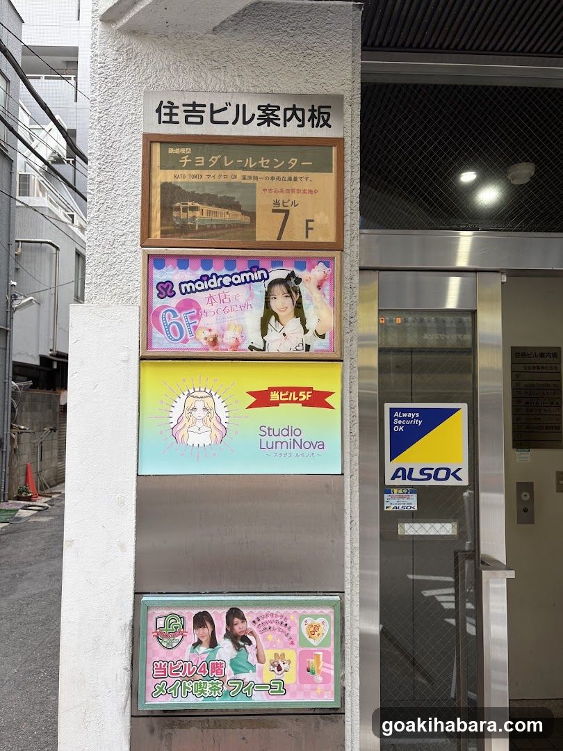 Photo of the exterior entrance to a building with multiple shop signs mounted on the wall. Visible signage includes 'チヨダールセンター' at 7F, 'maidreamin' at 6F, 'Studio LumiNova' at 5F, and 'メイド喫茶フィーユ' at 4F. A security sticker is affixed to the glass door.