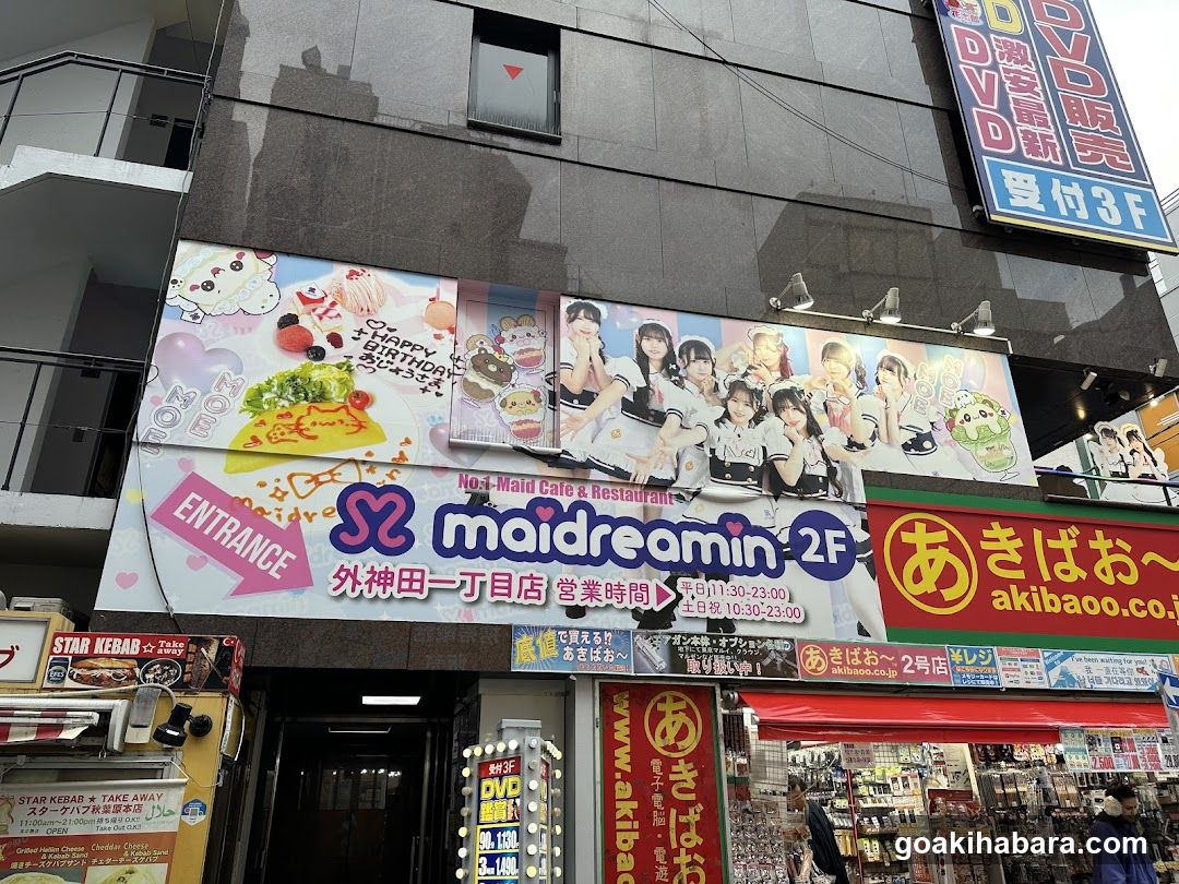 A photo of the physical exterior entrance and storefront facade featuring a large banner with anime-style characters, 'maidreamin' signage, and multiple shop signs including 'akibao', 'Star Kebab', and DVD rental ads.
