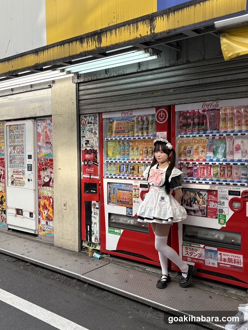 A photo of a street-level storefront featuring multiple red and white Coca-Cola vending machines lined up along the sidewalk. A young woman in a maid costume stands confidently in front of the machines, posing for the camera. The building facade above has yellow and blue paint with visible wear and tear.