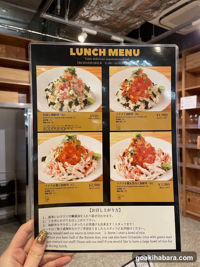 A hand holds a laminated lunch menu featuring four rice dishes with seafood toppings, including salmon roe and eel sauce, priced between ¥1,910 and ¥3,980. The menu includes Japanese text instructions for serving the meal.