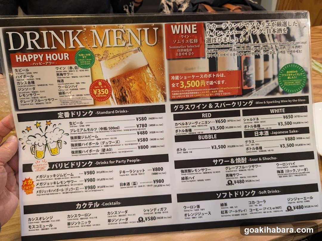 A close-up of a drink menu board featuring sections like Happy Hour, Standard Drinks, Sparkling Wine & Sake, Sour & Shochu, and Soft Drinks. The menu includes prices in yen and lists various beverages such as beer, wine, sake, cocktails, and soft drinks.