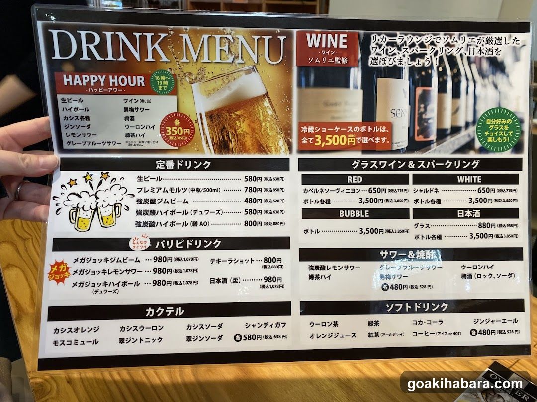 A close-up photo of a printed drink menu held by a hand, featuring sections like Happy Hour, Still Drinks, Glass Wine & Sparkling, and Soft Drinks.