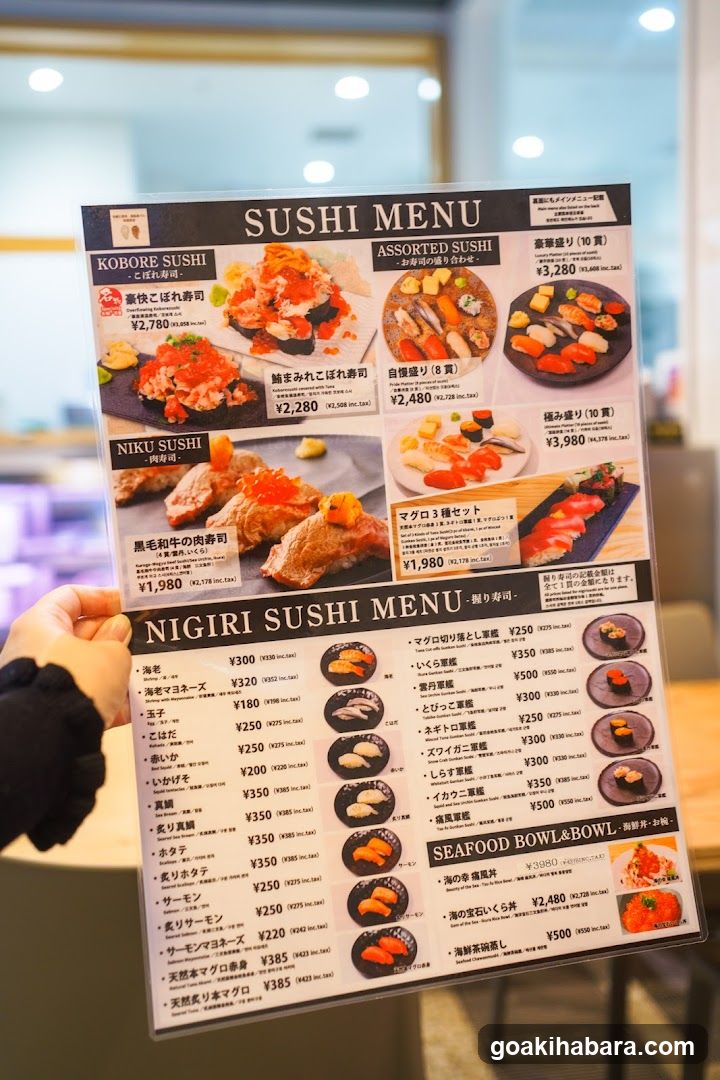 A person holds a sushi menu featuring sections like Kobore Sushi, Assorted Sushi, Niku Sushi, and Nigiri Sushi Menu with prices in yen.