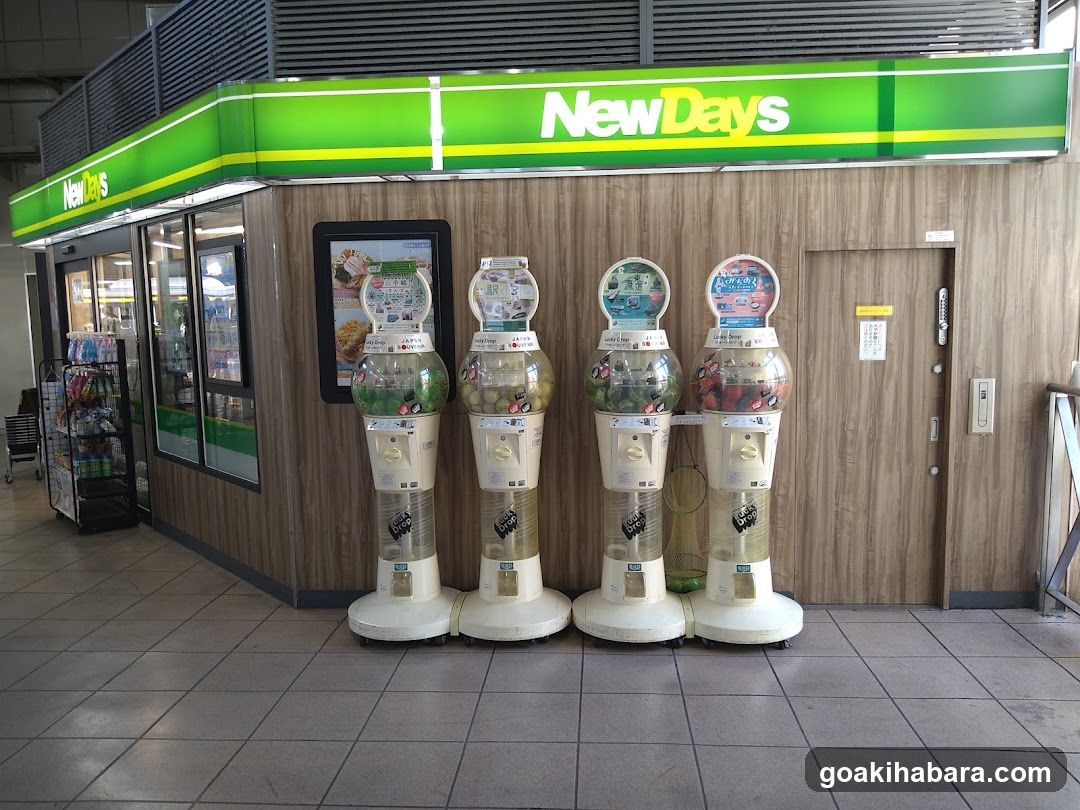 A NewDays convenience store entrance with four gumball machines lined up in front of the wooden facade. The store sign is visible above, and there are glass doors to the left showing the interior. A menu board is mounted on the wall near the entrance.