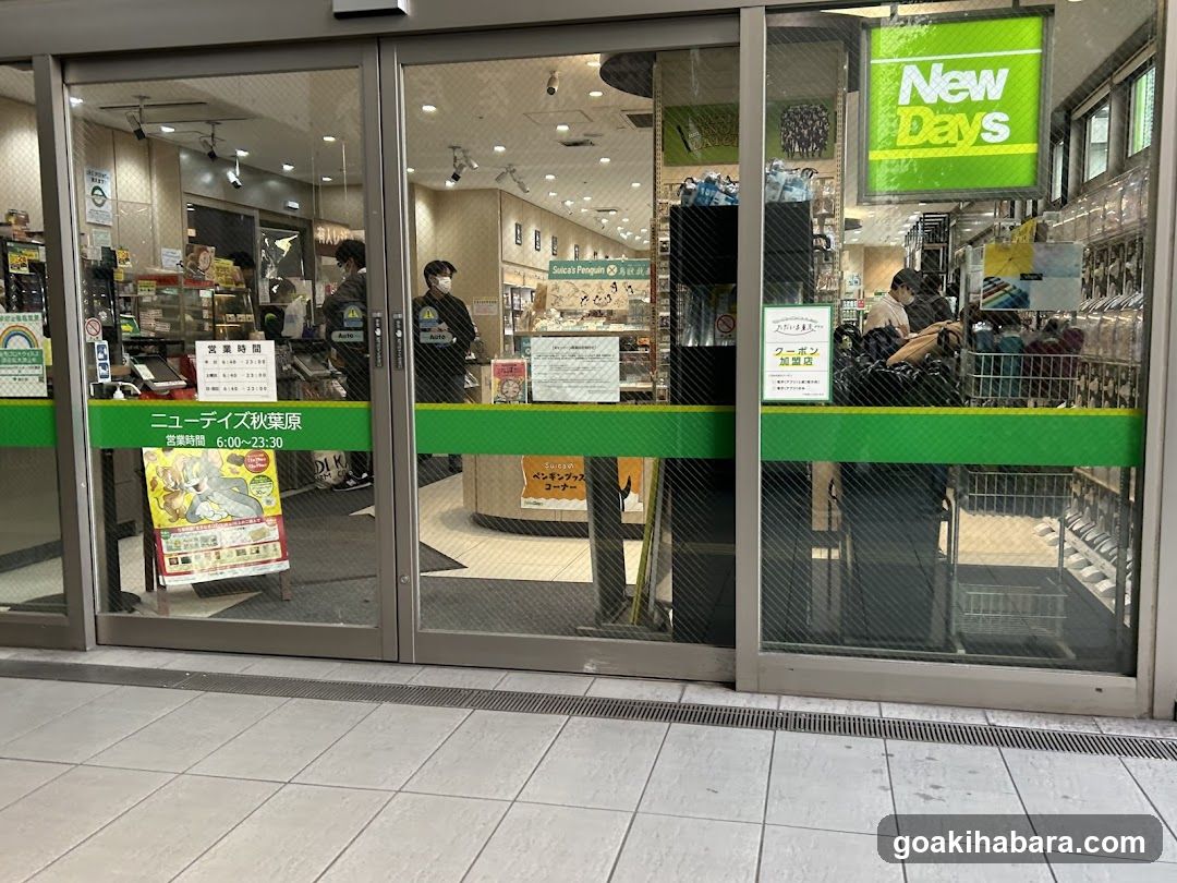 Photo of the physical exterior entrance of a New Days convenience store, featuring glass sliding doors with green branding and signage indicating business hours from 6:00 to 23:30. Inside, shelves stocked with products are visible through the transparent doors.