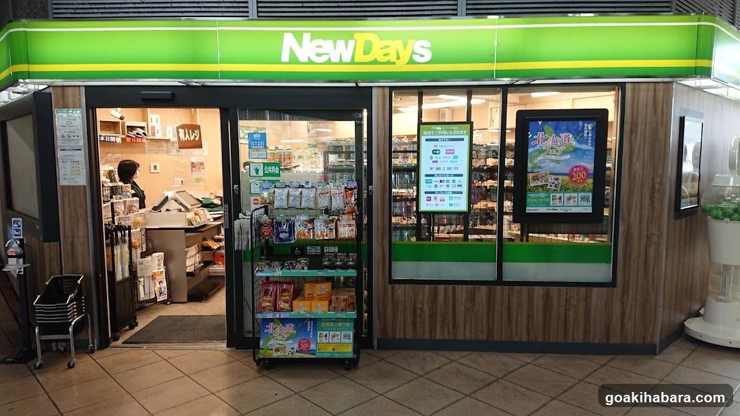 The photo shows the exterior entrance of a NewDays convenience store, featuring its distinctive green and yellow signage above the glass doors. Inside, shelves are stocked with various products, and there is a promotional display near the entrance.
